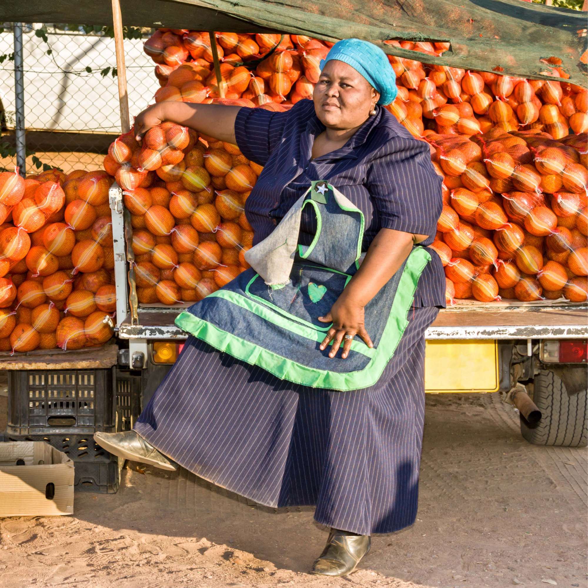 Photograph of a woman sitting on the back of a truck loaded with numerous orange pumpkins, suggesting a market or harvest setting. She wears a blue dress, green and blue apron, and blue headscarf, with pumpkins stacked behind her and crates visible underneath the truck.