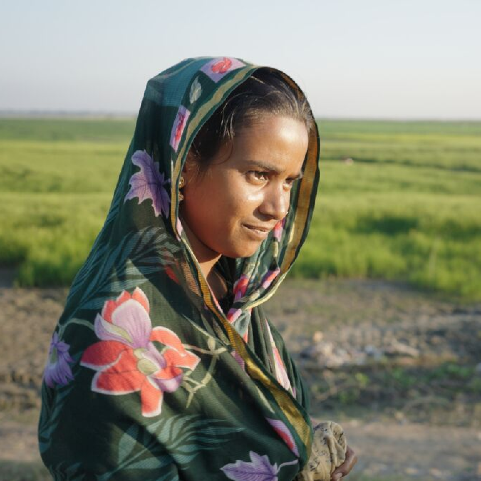 Photograph of a young woman from Bangladesh wearing a dark green shawl with large pink and purple floral patterns, standing outdoors near a field with green crops under a clear sky. The scene highlights rural or agricultural setting with natural lighting and a blurred background emphasizing the subject.