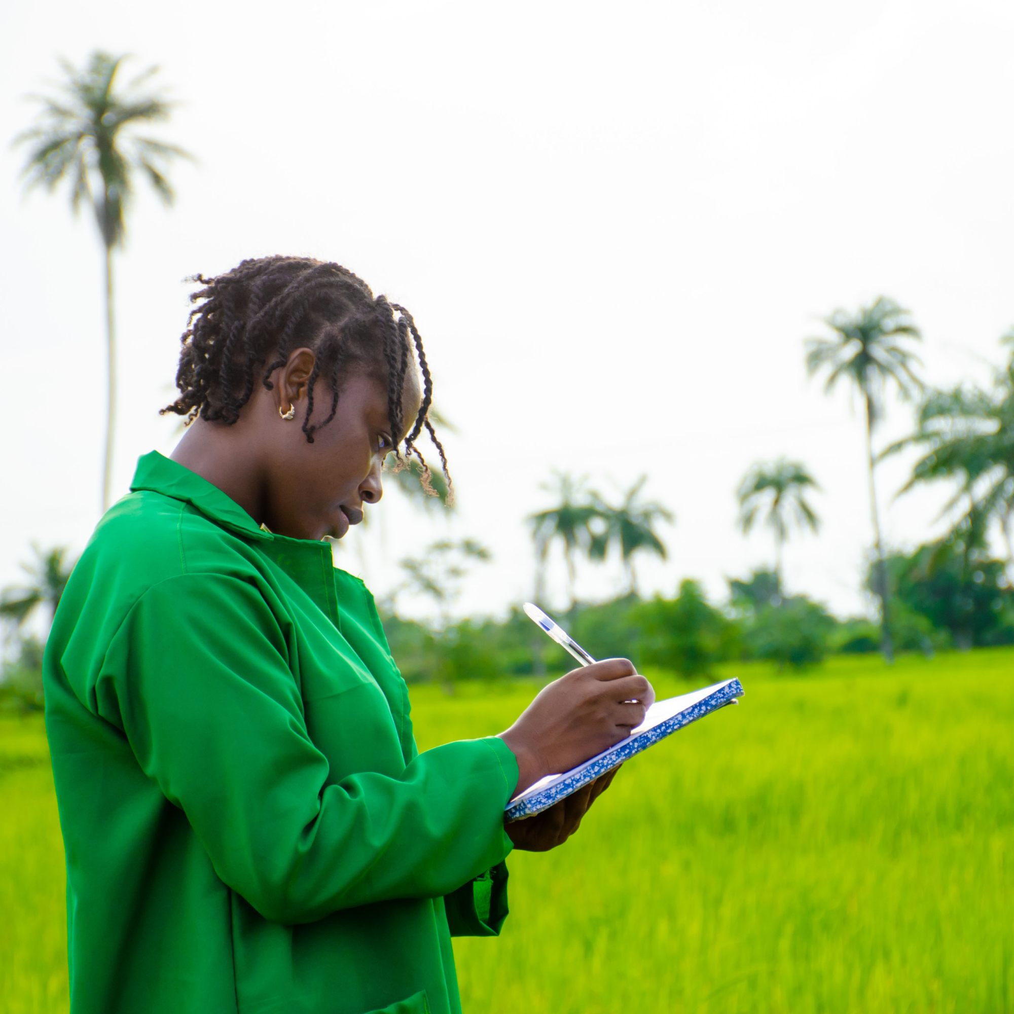Photograph of a woman wearing a green jacket standing in a lush green field, writing in a notebook with a pen. Background includes tall palm trees and a cloudy sky, suggesting an outdoor agricultural or environmental study setting.
