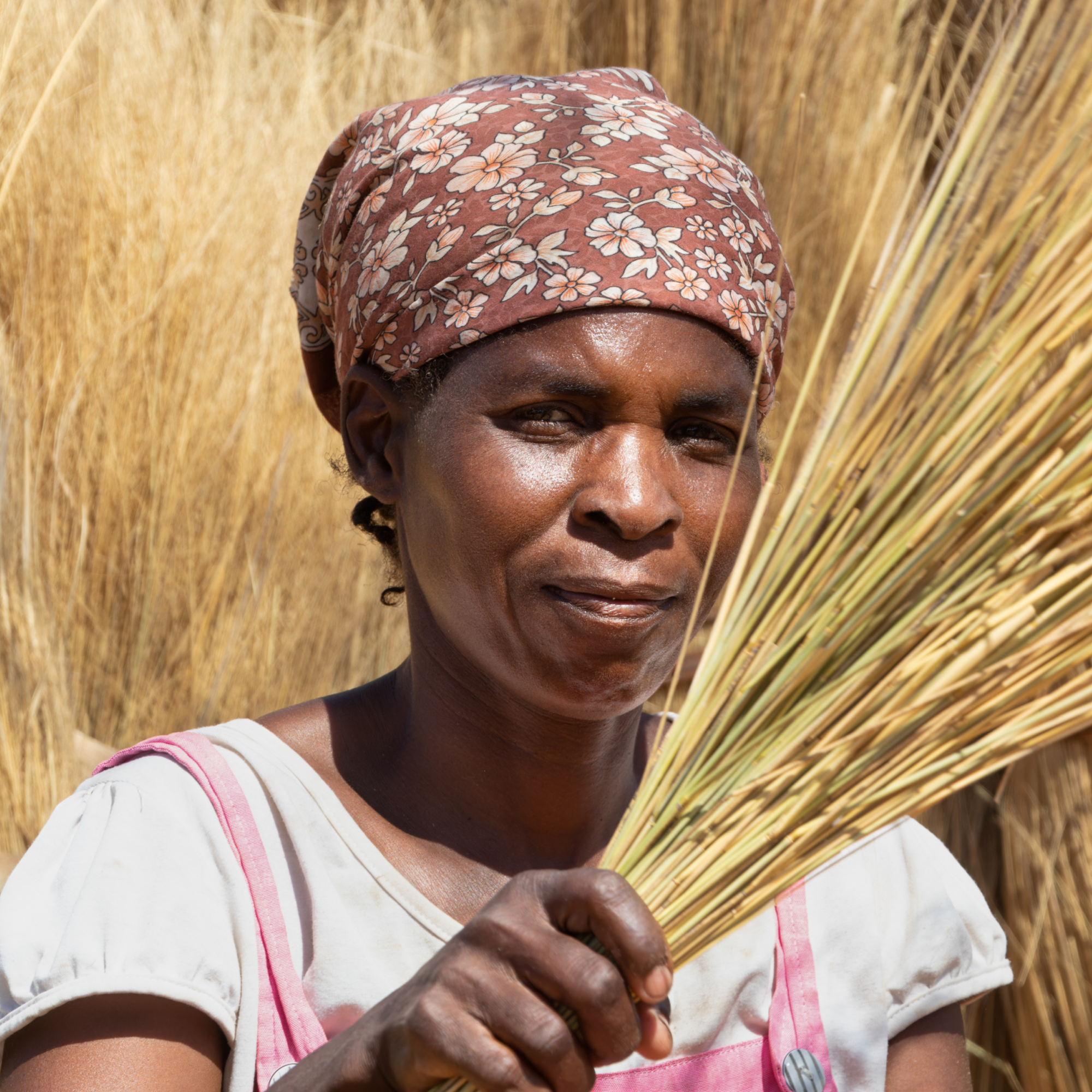 Photograph of a person wearing a floral headscarf and white shirt with pink suspenders, holding a bundle of dried grass or straw. Background filled with similar dried grass, suggesting an agricultural or rural setting.