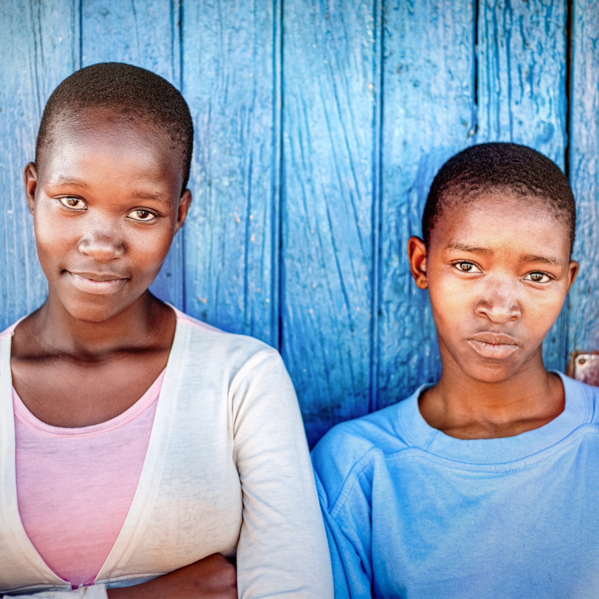 Photograph of two young people standing side by side against a textured blue wooden wall. Both wear casual clothing with one in a light pink top and white cardigan, the other in a light blue shirt, highlighting a contrast in attire and posture.