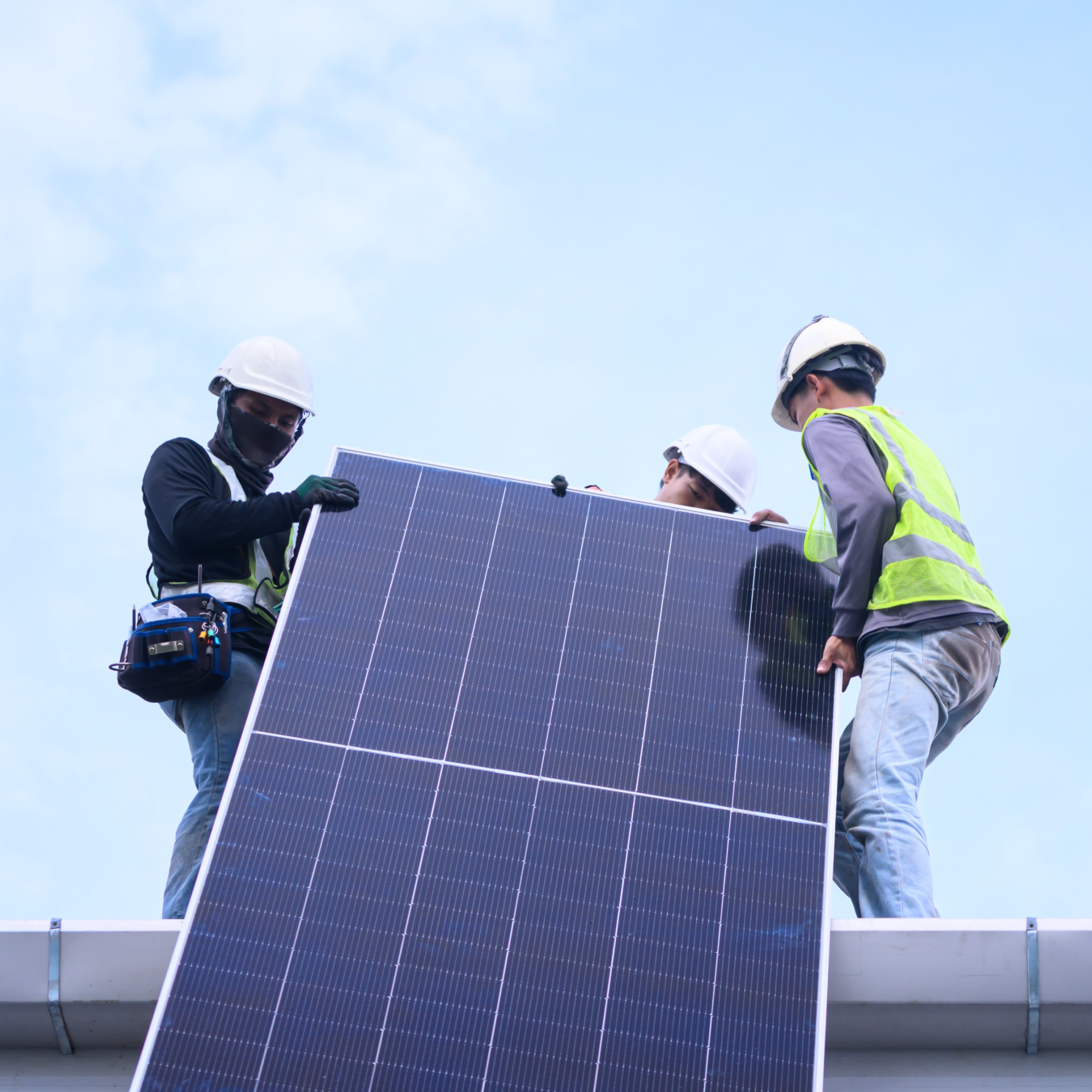 Photograph showing three workers wearing safety helmets and one in a reflective vest installing a large solar panel on a rooftop under a clear sky. The scene highlights teamwork and safety measures during solar panel installation for renewable energy purposes. (Project FP009)