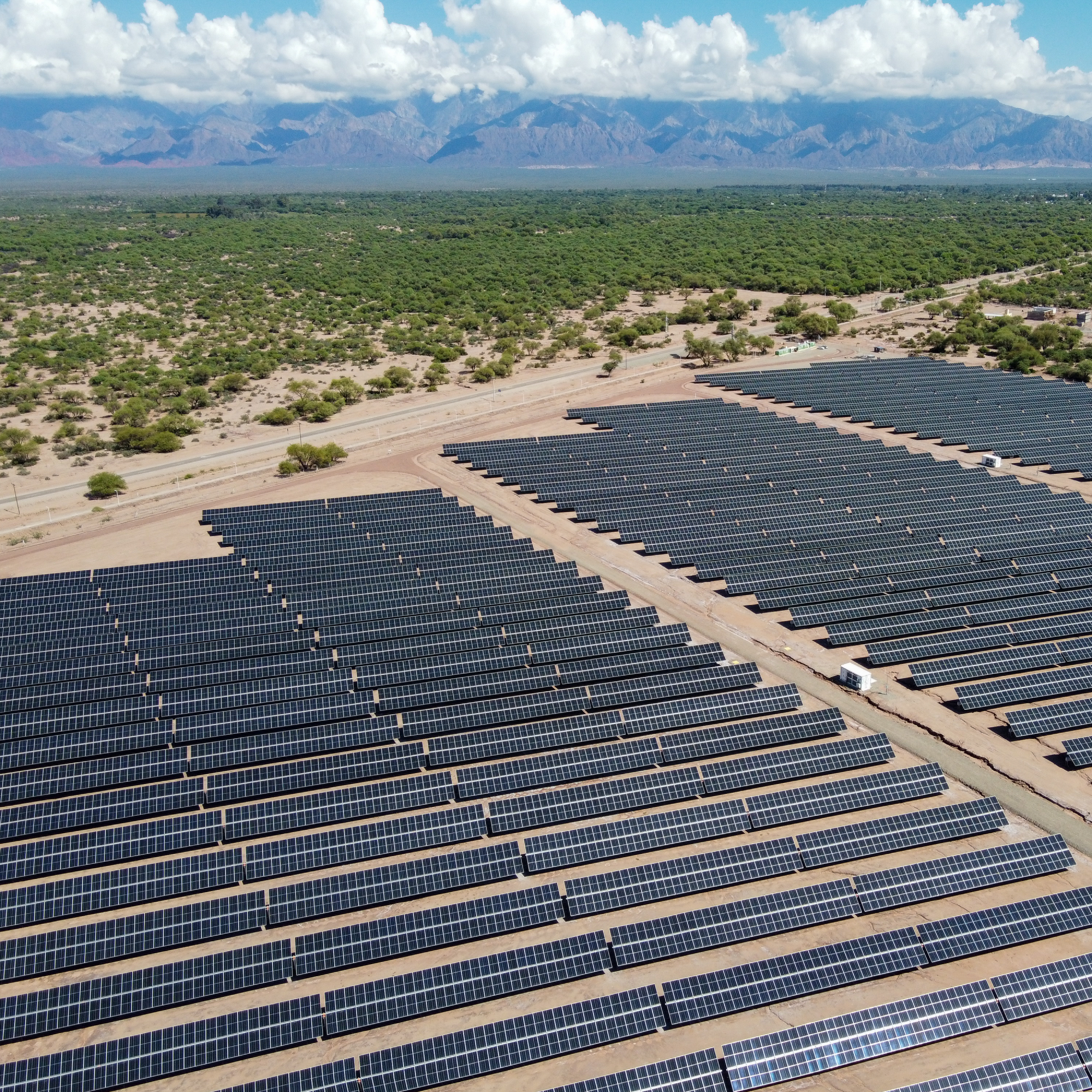 Aerial photograph of a large solar farm with numerous rows of dark blue solar panels arranged in parallel lines across a desert landscape. Green vegetation surrounds the solar farm, with mountains and a partly cloudy sky visible in the background, highlighting renewable energy infrastructure in a natural setting. (Project FP030)
