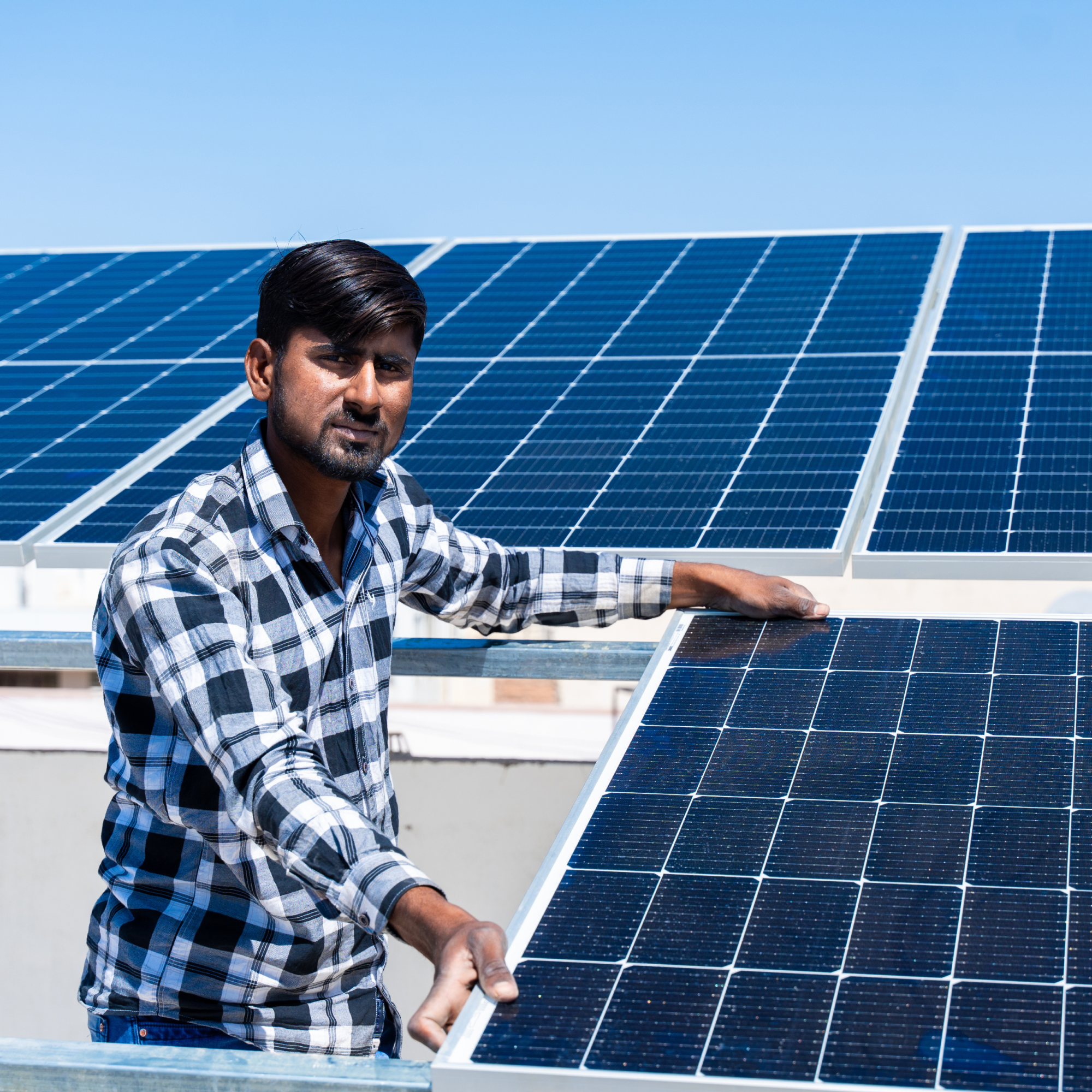 Photograph of an Indian man wearing a black-and-white checkered shirt installing or inspecting blue solar panels on a rooftop under a clear blue sky. The scene highlights renewable energy technology with multiple solar panels arranged in rows, emphasizing solar power generation. (Project FP081)