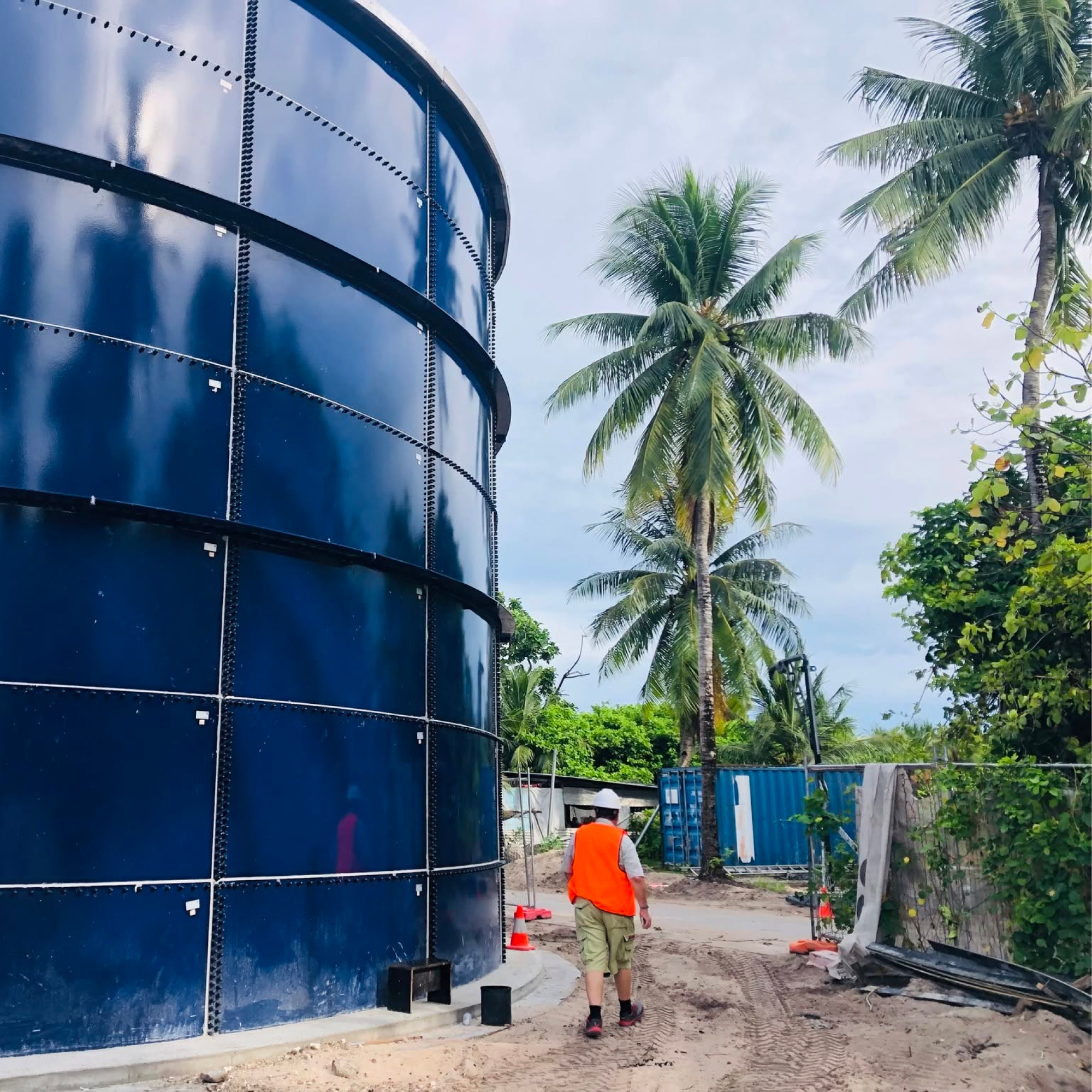 Photograph of a large, dark blue cylindrical water tank next to a dirt path with a person wearing an orange safety vest and hat walking away from the camera. Surrounding elements include tall palm trees, green foliage, construction cones, and a blue shipping container, indicating an outdoor industrial or construction site. (project FP091)