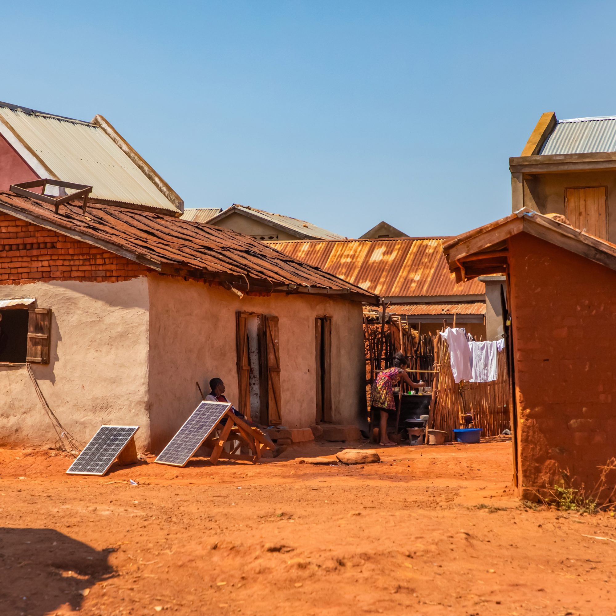 Photograph of a rural homestead with multiple small buildings featuring metal and tiled roofs under a clear blue sky. Two solar panels lean against a mud-walled house, while a person washes clothes near a wooden fence with laundry hanging, highlighting use of solar energy and daily life activities. (Project FP093)
