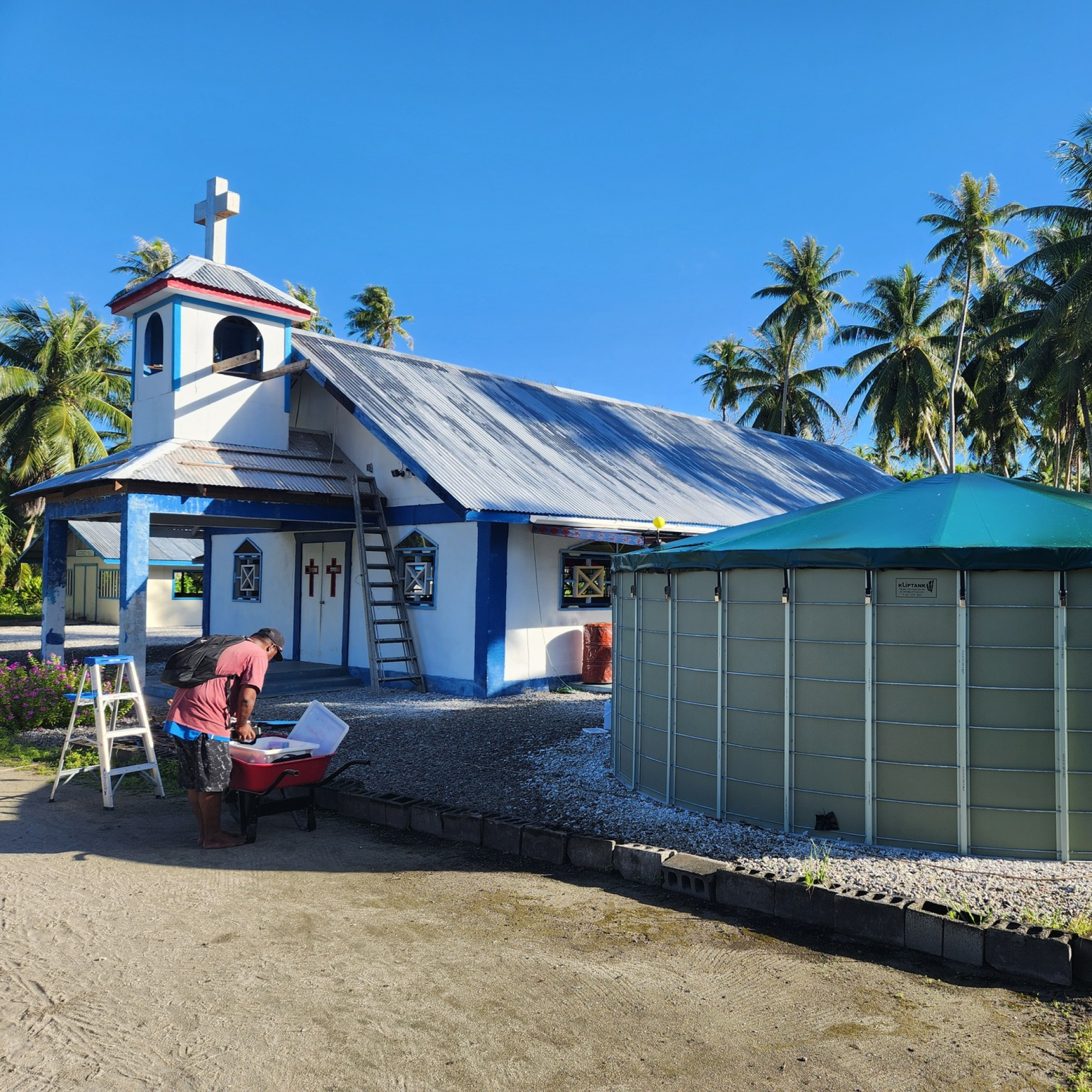 Photograph of a small church with white walls and blue trim, featuring a cross on top and palm trees in the background under a clear blue sky. A person is working near a red wheelbarrow beside a large green water tank, with a ladder leaning against the church roof, suggesting maintenance or preparation activities. (Project FP112)