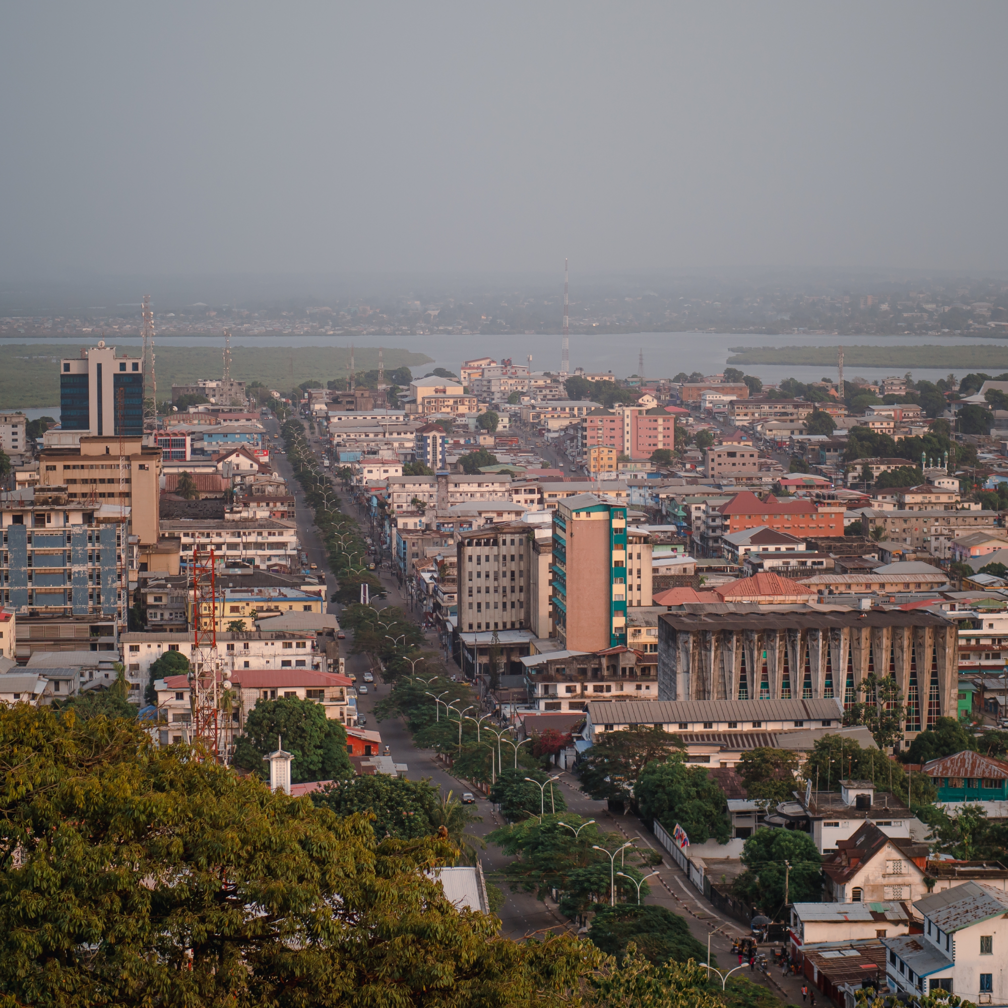 Photograph of an urban cityscape featuring a central road lined with buildings of varying heights and architectural styles, extending toward a body of water in the background. The scene includes greenery in the foreground, several tall communication towers, and a mix of commercial and residential structures under a hazy sky. (project FP160)
