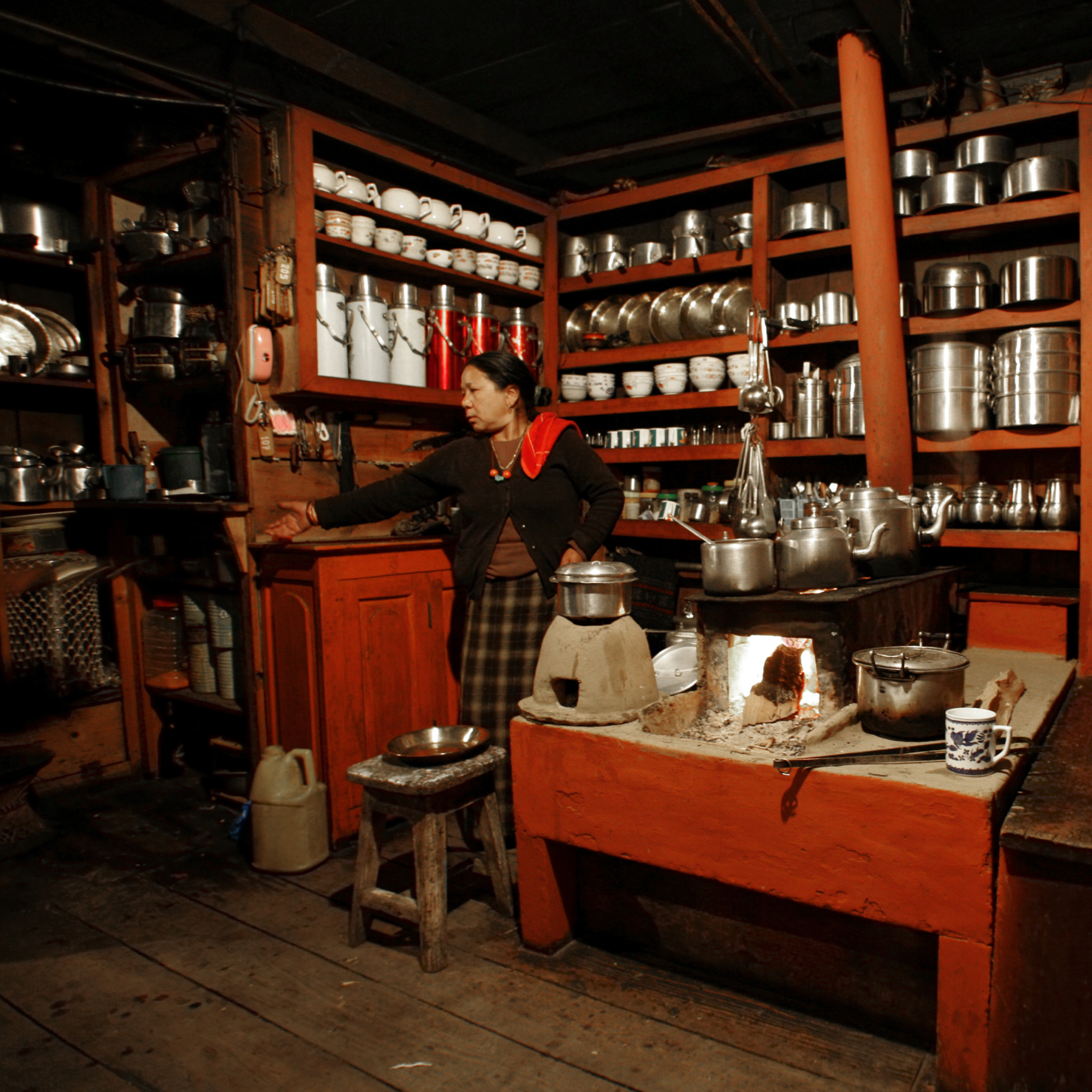 Photograph of a traditional kitchen featuring wooden shelves stocked with metal pots, pans, and white ceramic cups, centered around a clay stove with a fire burning beneath cooking vessels. A woman stands near a wooden cabinet, highlighting rustic cooking setup with warm lighting and various utensils arranged neatly on shelves. (project FP172)