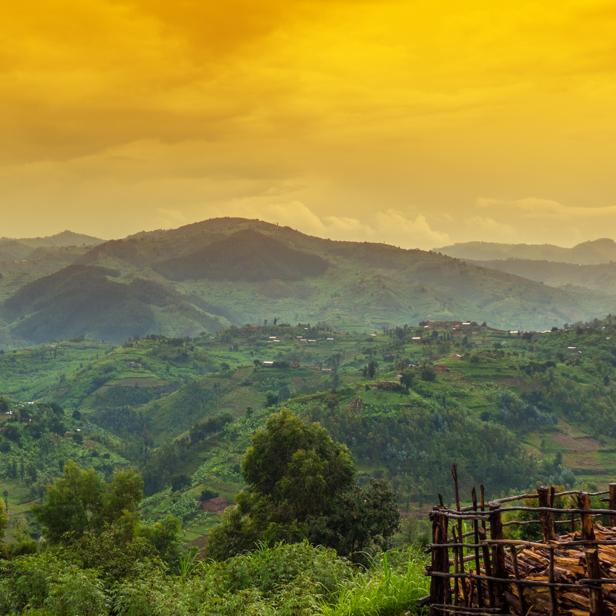 Photograph of a lush green mountainous landscape under a vibrant yellow-orange sky during sunset or sunrise. Foreground features dense vegetation and a rustic wooden fence, while rolling hills and scattered small buildings extend into the misty distance. (project FP217)