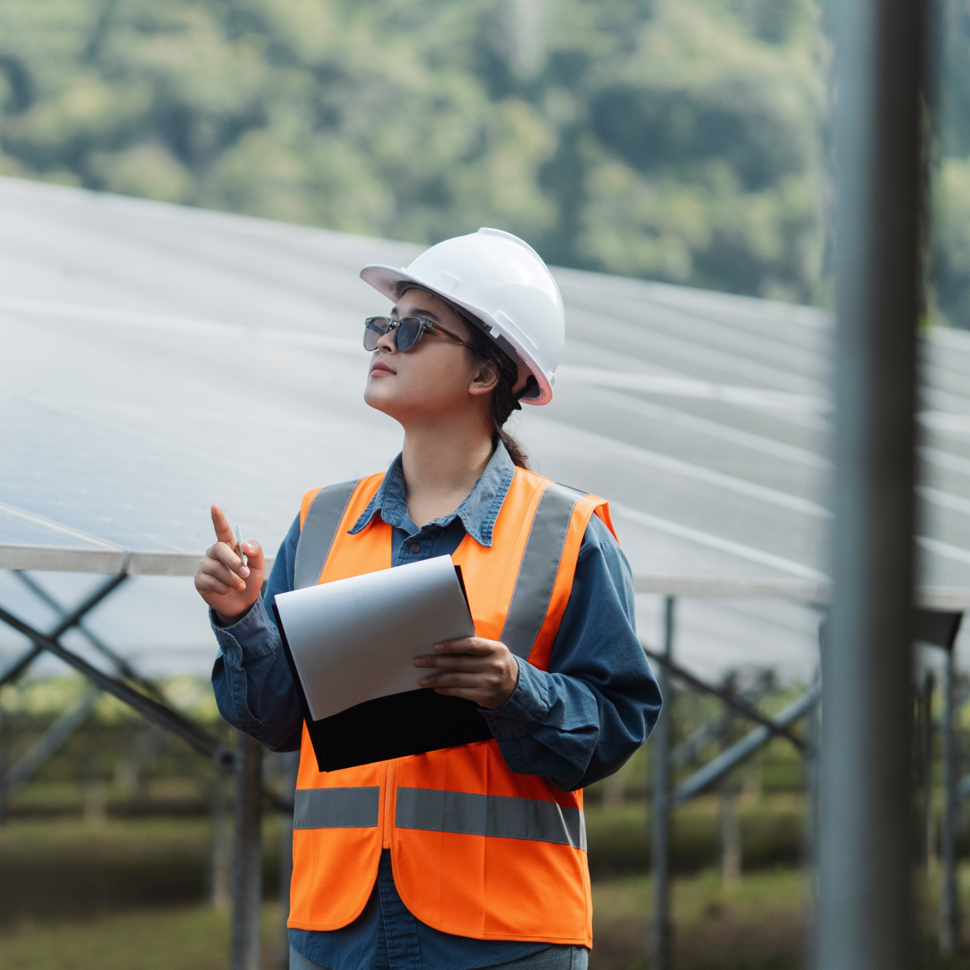 Photograph of a person wearing a white hard hat and orange safety vest holding a clipboard and pointing upward at a solar panel installation. The setting highlights renewable energy infrastructure with rows of solar panels and a blurred natural background. (project FP240)