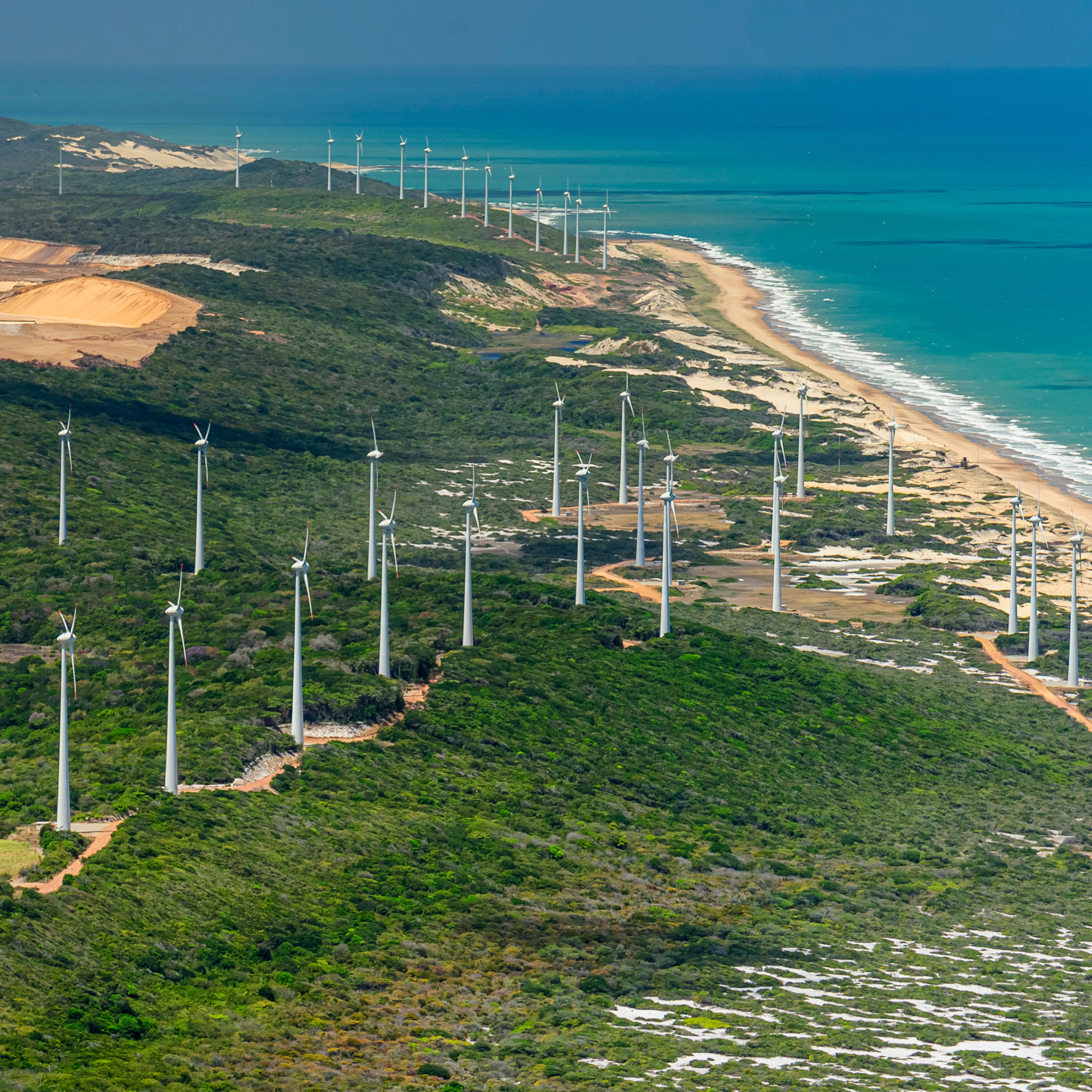 Aerial photograph of a coastal wind farm featuring numerous white wind turbines arranged in rows along green vegetation near a sandy beach with turquoise ocean waters. The image highlights renewable energy infrastructure integrated into a natural landscape, showing turbines spaced evenly with access roads visible between them. (project FP285)