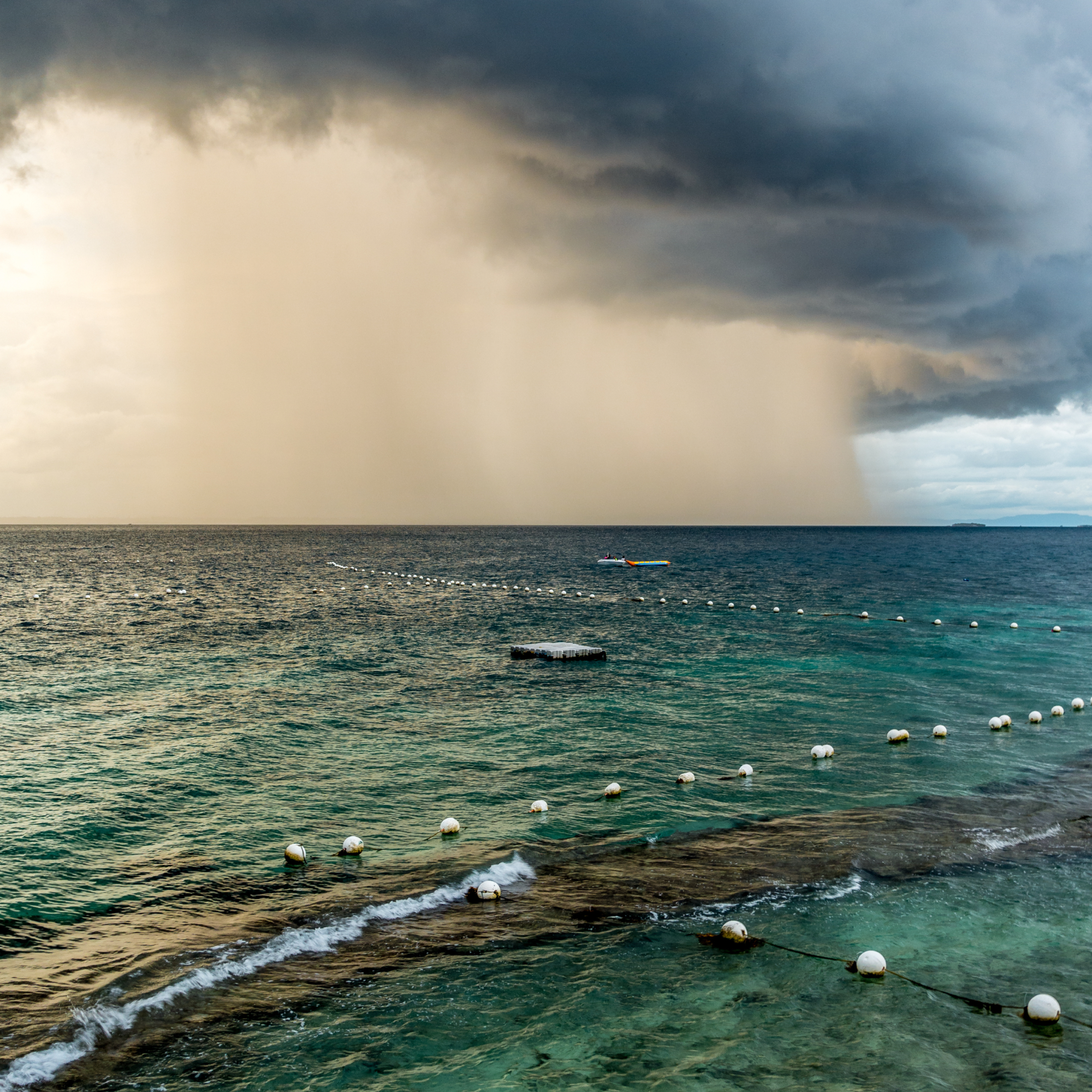 Photograph showing a storm over a body of water with dark clouds and heavy rain in the distance. Foreground features calm, clear water with white buoys marking a swimming area and a floating platform. (Project SAP010)