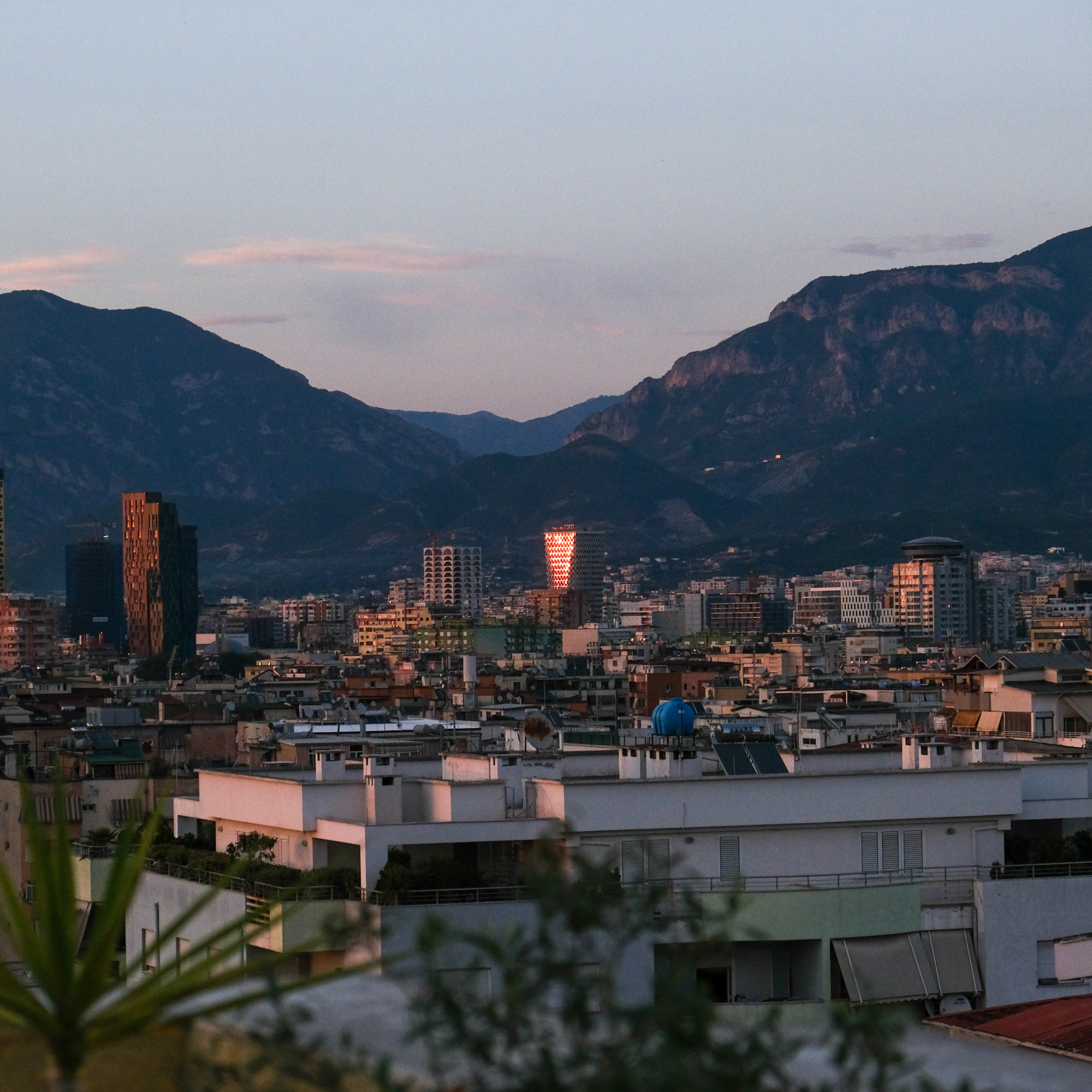 Photograph of a cityscape at dusk with mountains in the background, showcasing a mix of residential and commercial buildings. A tall building with reflective glass panels illuminated by sunlight stands out centrally, contrasting with surrounding structures under a soft purple and blue sky. (Project SAP041)