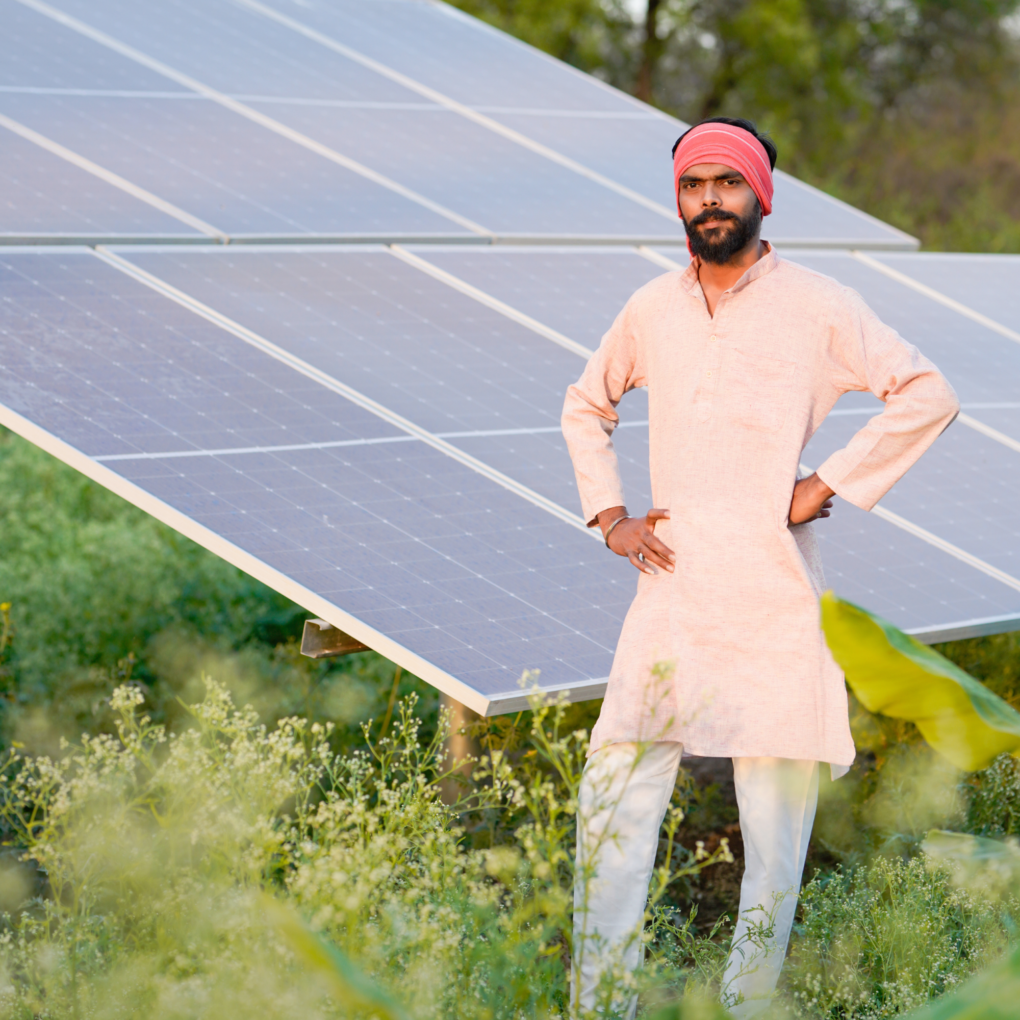 Photograph showing a man standing confidently with hands on hips next to a large solar panel array in a green outdoor setting. The scene highlights renewable energy adoption in rural or agricultural areas, with the man wearing traditional attire and the solar panels featuring a grid of blue photovoltaic cells. (Project SAP064)