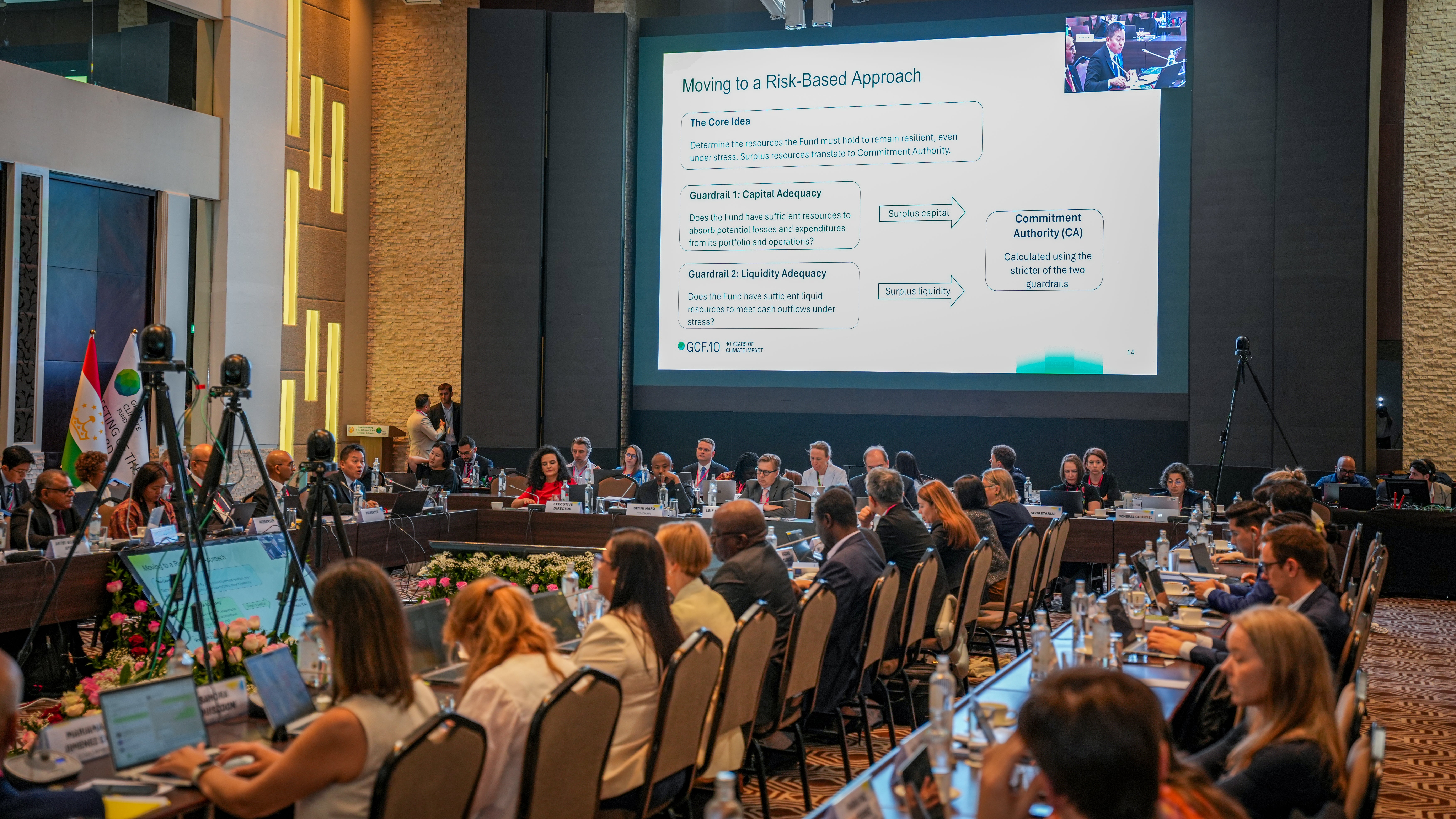 Photograph of a large conference room with numerous attendees seated around a U-shaped table, focusing on a presentation displayed on a large screen titled "Moving to a Risk-Based Approach."