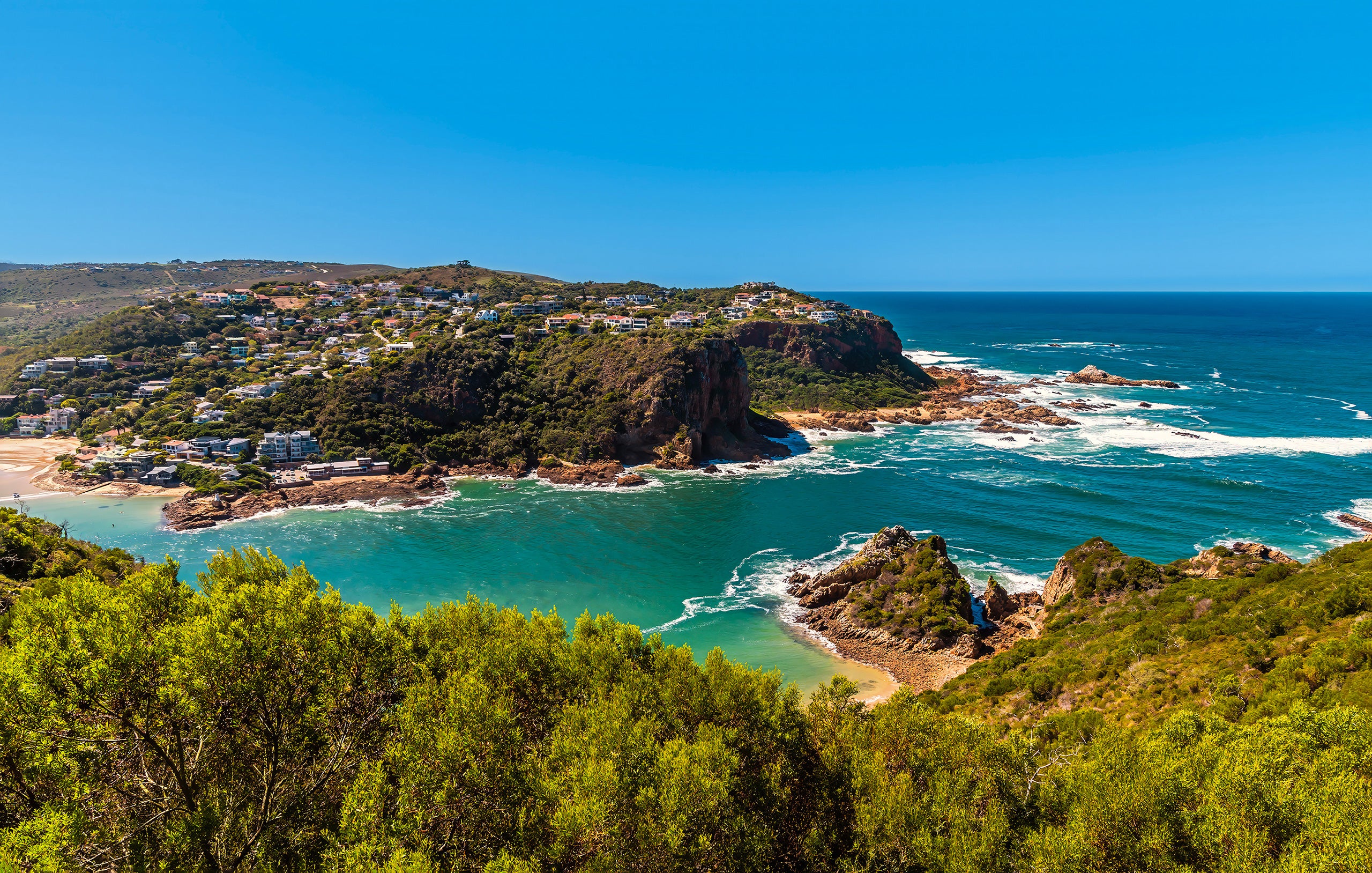 Rocky coastal headland covered in green vegetation and hillside homes overlooking turquoise bays and waves breaking along the shoreline under a clear blue sky.