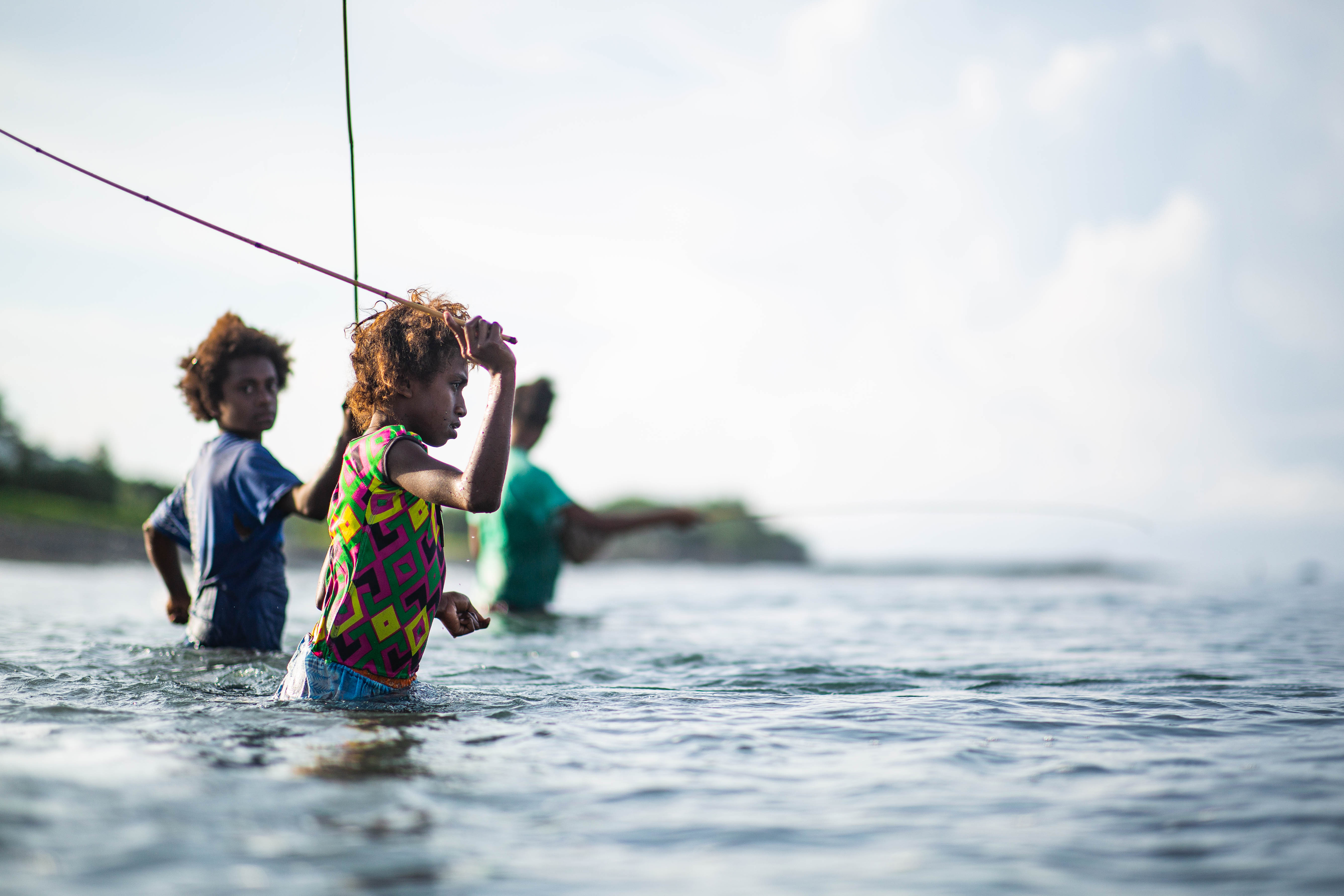 Photograph of three children standing waist-deep in water, holding fishing lines, likely engaged in fishing activity. The scene features a calm body of water with a blurred shoreline and cloudy sky in the background, highlighting a peaceful outdoor moment.