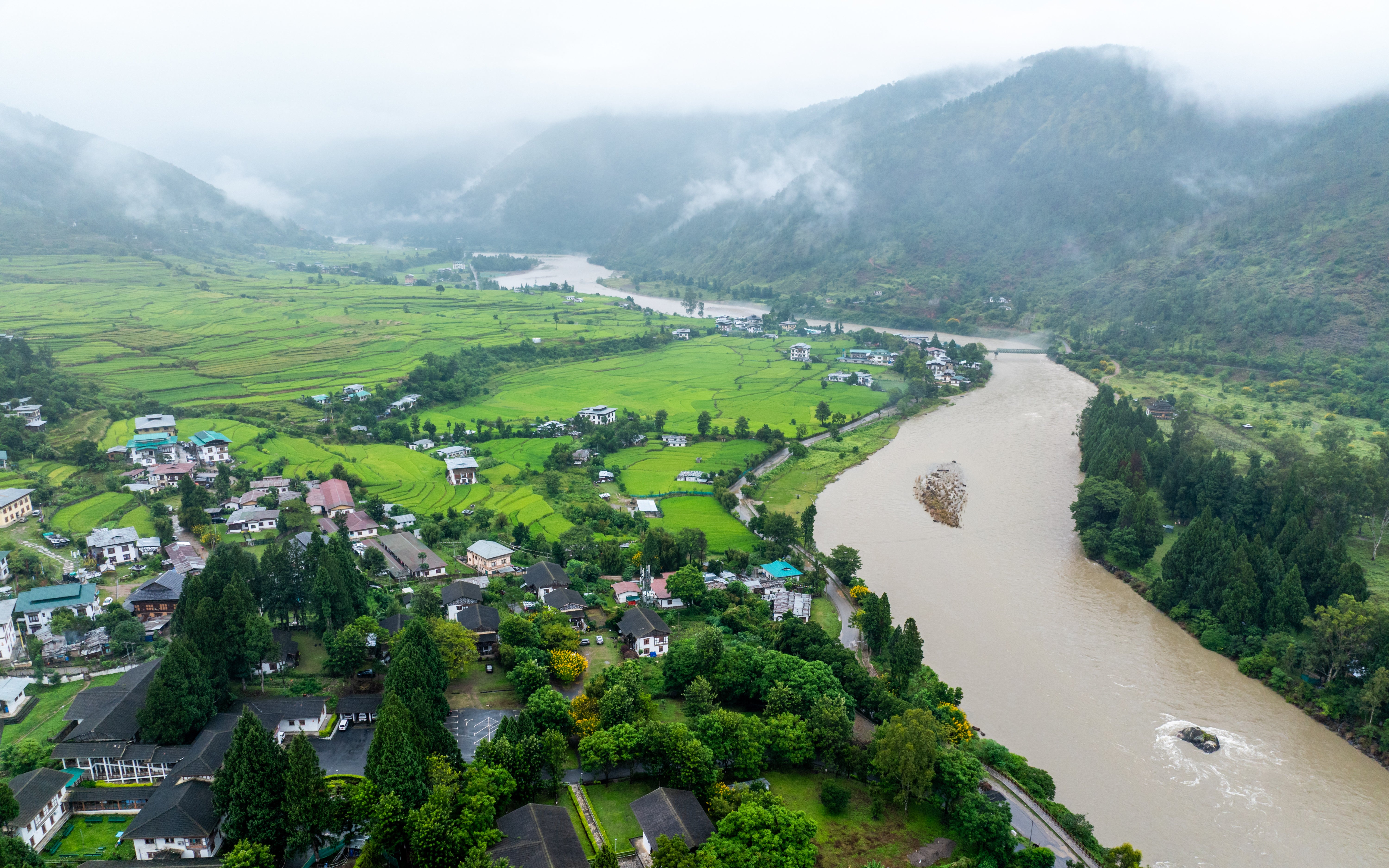 Aerial view of a winding river through a lush mountain valley with farmland, villages and mist-covered hills.