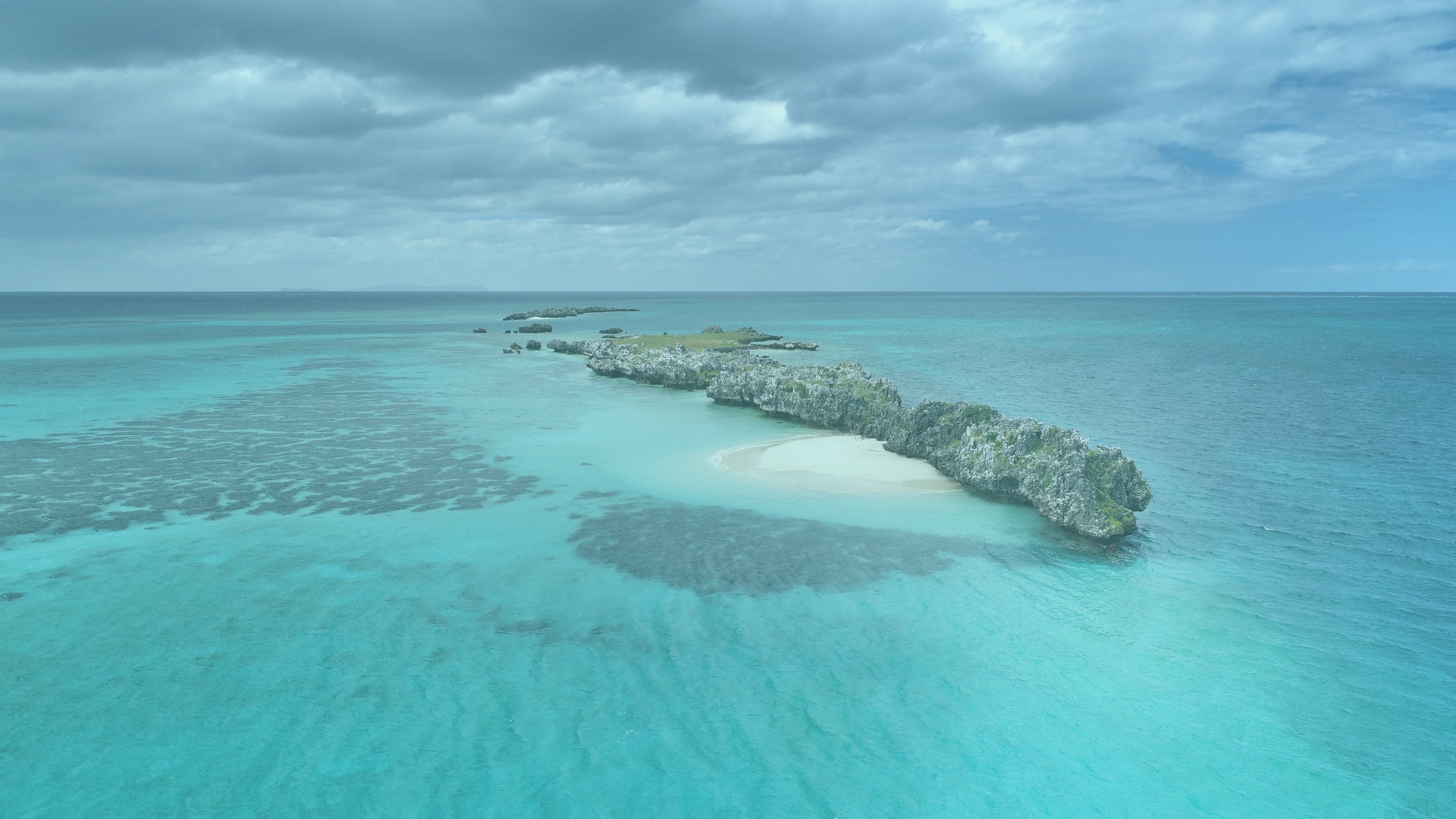 Aerial view of a rocky islet and white sandbar surrounded by shallow turquoise ocean.