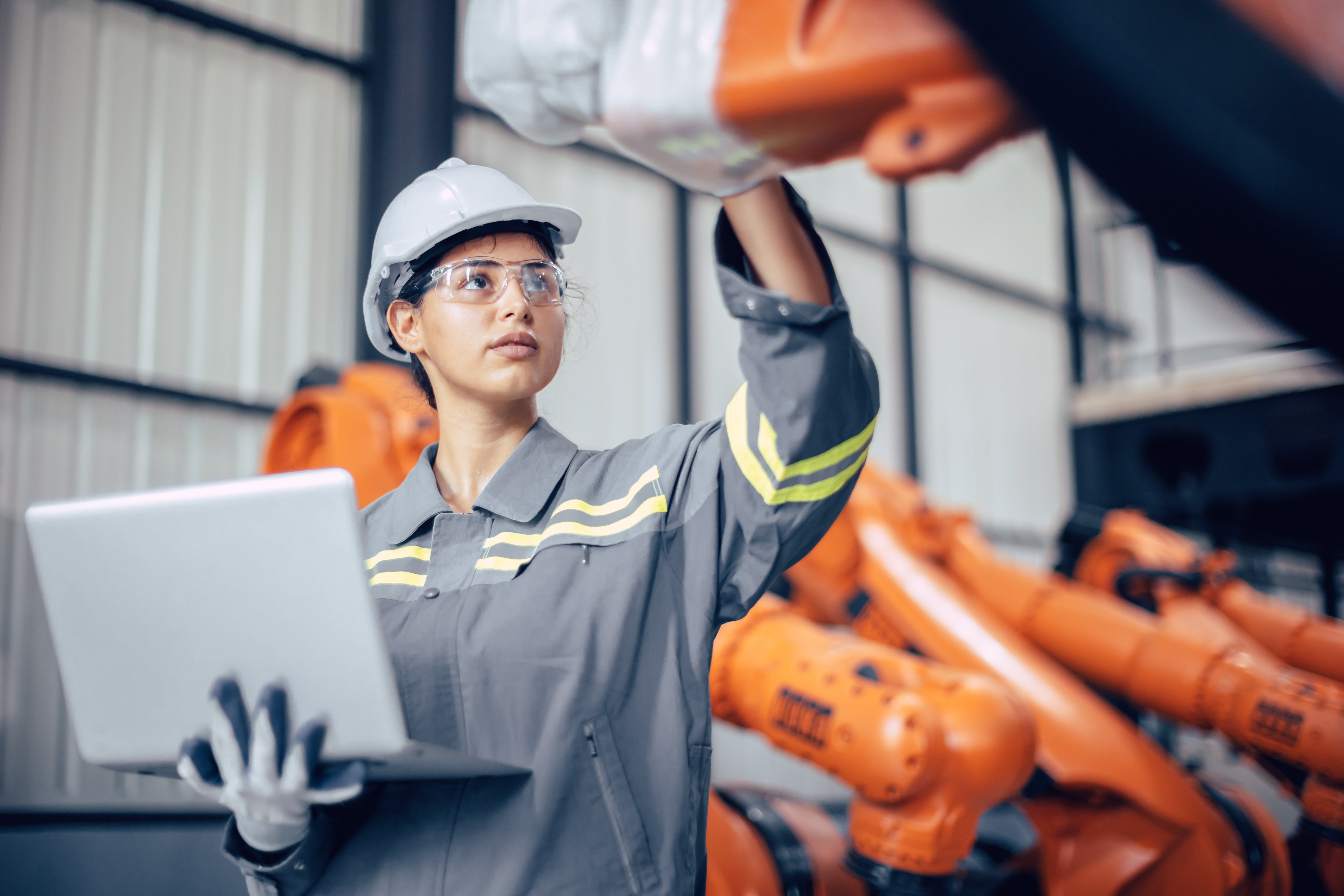 Worker wearing a hard hat and safety glasses inspecting industrial equipment while holding a laptop.