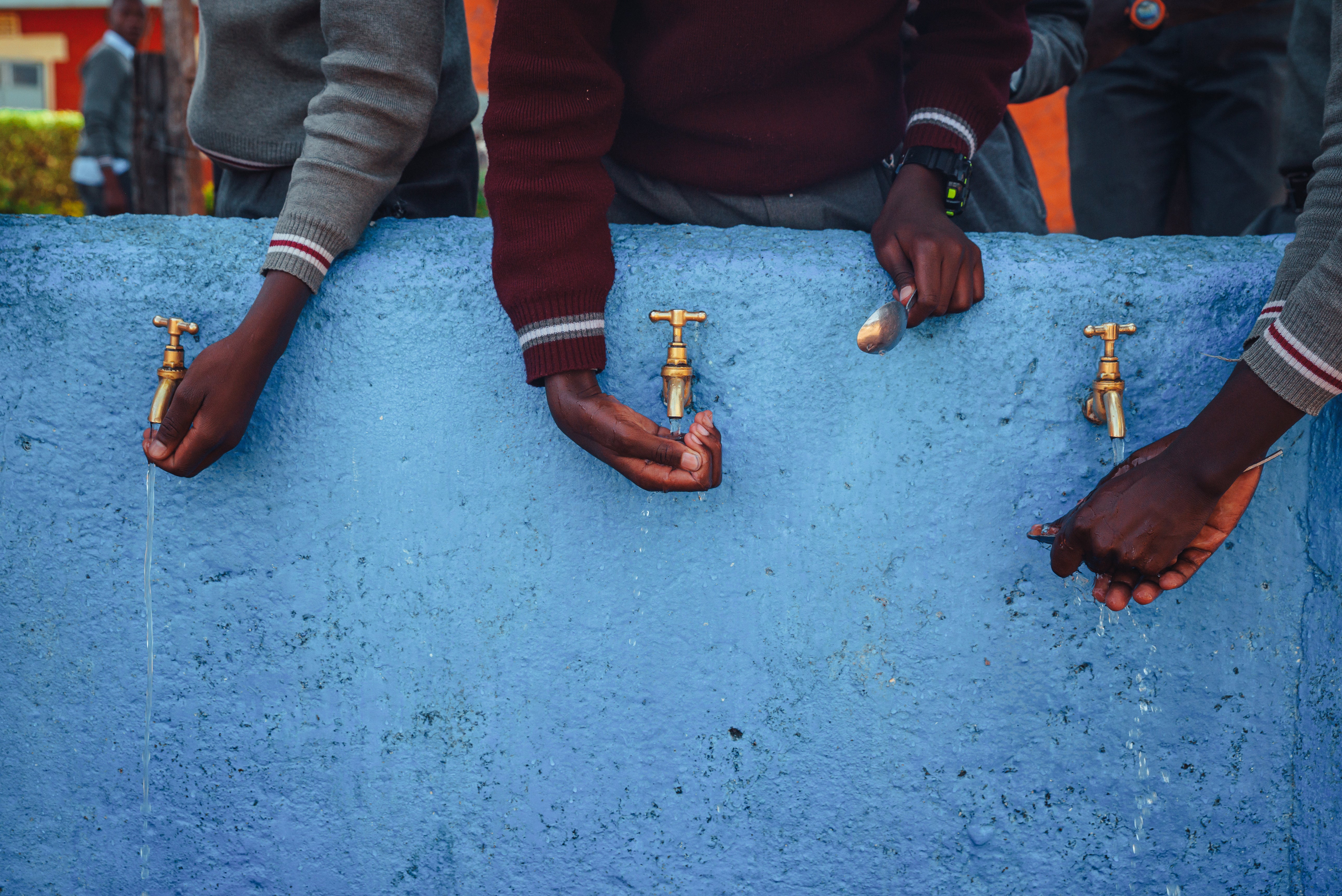Students washing their hands at outdoor taps above a blue concrete basin.